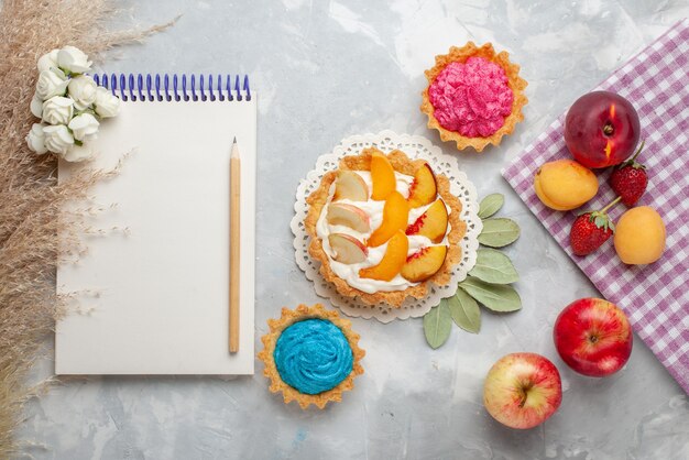 Vista superior de un pequeño pastel cremoso con frutas en rodajas y crema blanca junto con pasteles cremosos y frutas en un piso blanco claro, pastel de frutas, galleta dulce