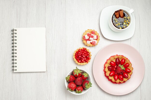 Vista superior pequeño pastel cremoso con frutas y taza de té en el escritorio blanco