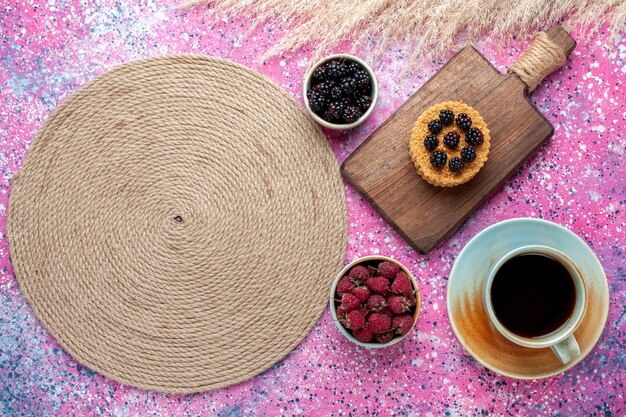 Vista superior del pequeño pastel con diferentes bayas y taza de té en superficie rosa