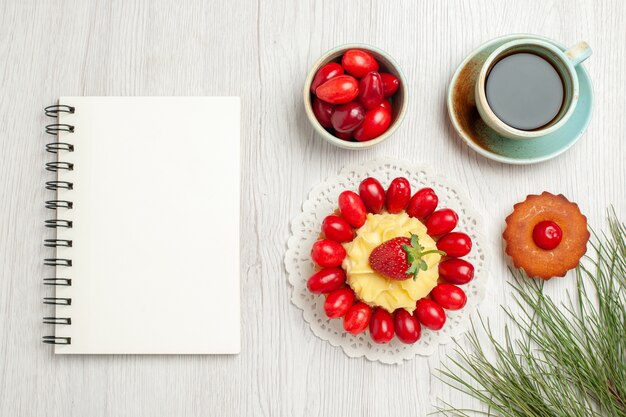 Vista superior del pequeño pastel con frutas y una taza de té en el escritorio blanco