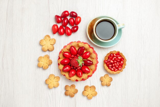 Vista superior del pequeño pastel con galletas de frutas y una taza de té en el escritorio blanco