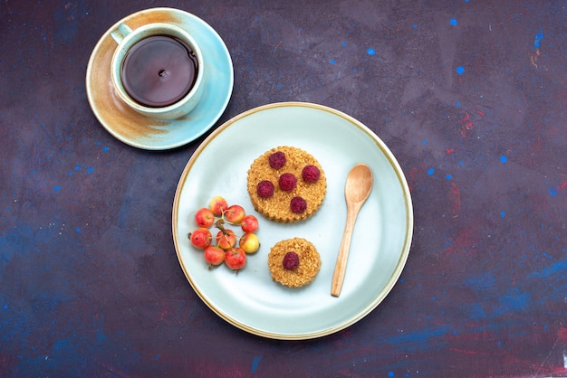 Vista superior del pequeño pastel redondo con frambuesas frescas dentro de la placa con té en la superficie oscura