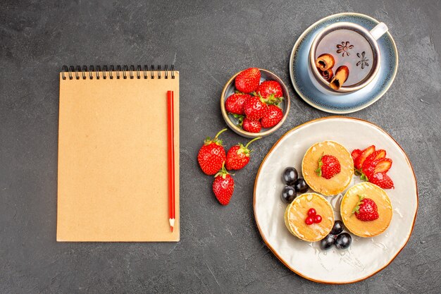 Vista superior pequeños panqueques deliciosos con frutas y una taza de té en el piso gris pastel pastel de frutas