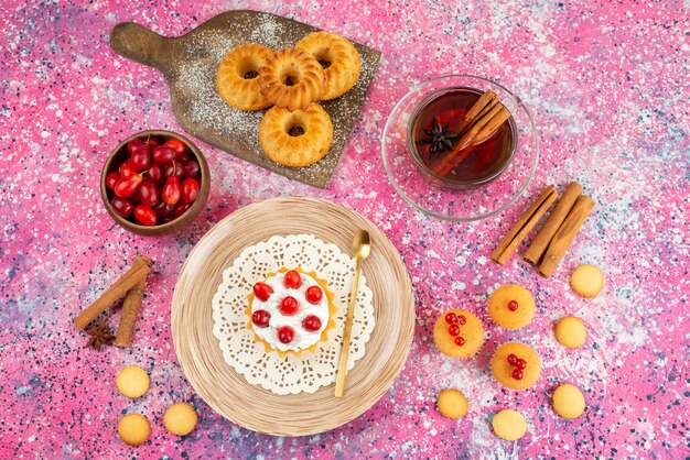 Vista superior de pequeños pasteles con crema fresca y frutas frescas junto con canela y té en la galleta de escritorio brillante