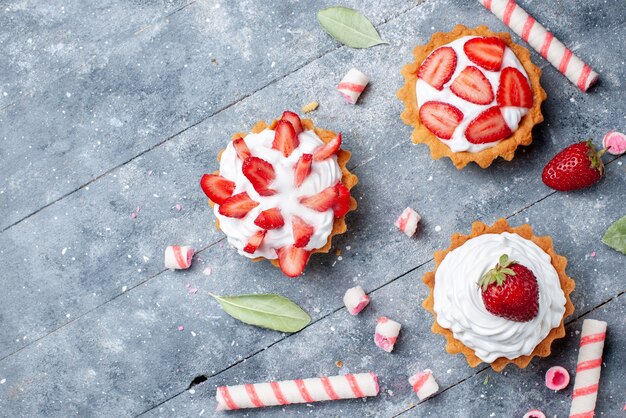 Vista superior de pequeños pasteles cremosos con fresas en rodajas y frescas junto con caramelos en el piso gris pastel de frutas dulce hornear baya
