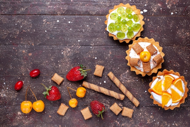 Vista superior de pequeños pasteles cremosos con rodajas de uvas naranjas junto con fresas en el escritorio de madera marrón pastel galleta fruta suave