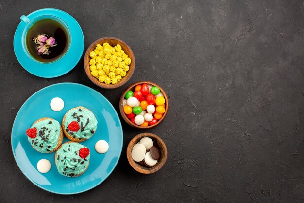 Vista superior de pequeños pasteles cremosos con una taza de té y caramelos en la superficie oscura, té, crema, pastel, galleta, postre color