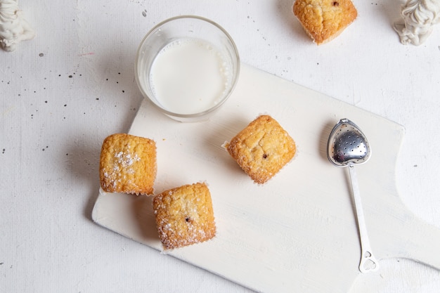 Una vista superior pequeños pasteles deliciosos con azúcar en polvo y un vaso de leche en la superficie blanca