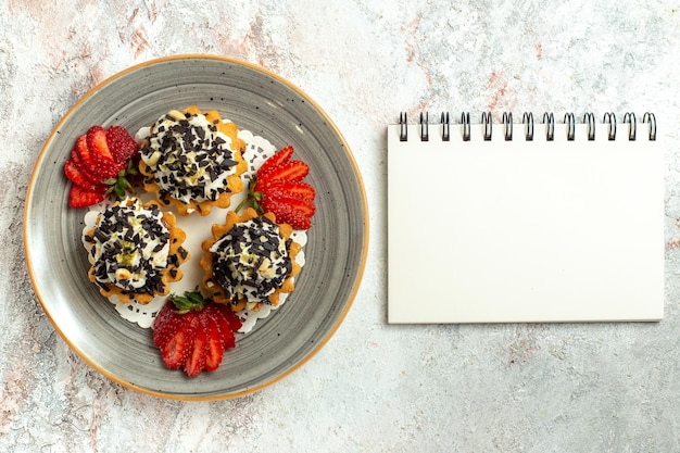 Vista superior pequeños pasteles deliciosos con crema y fresas en la superficie blanca torta de celebración de cumpleaños galleta dulce