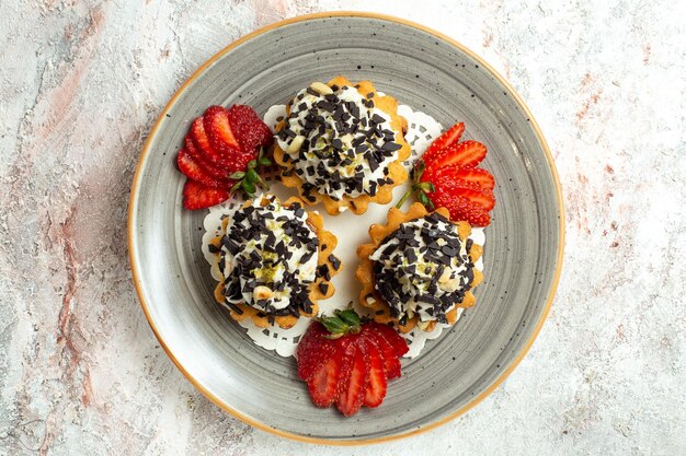 Vista superior pequeños pasteles deliciosos con crema y fresas en la superficie blanca torta de celebración de cumpleaños galleta dulce