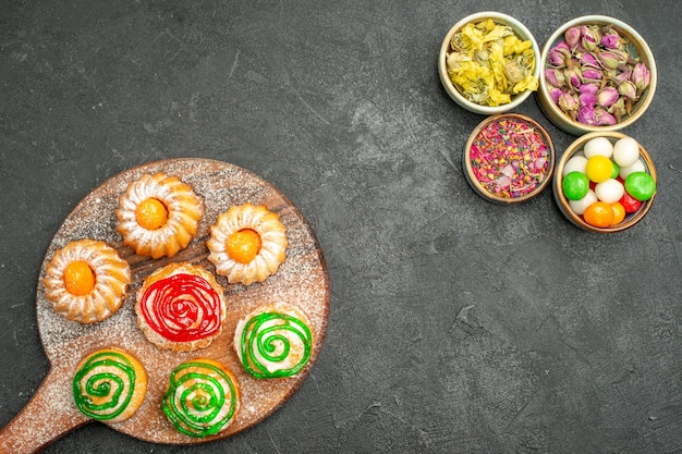 Vista superior de pequeños pasteles deliciosos con dulces y flores en la oscuridad