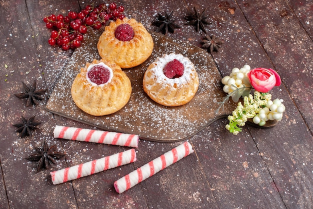 Vista superior de pequeños pasteles deliciosos con frambuesas y arándanos junto con caramelos de palo en el escritorio de madera pastel de frutas dulces hornear galletas baya