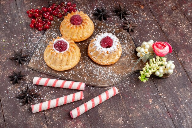 Vista superior de pequeños pasteles deliciosos con frambuesas y arándanos junto con caramelos de palo en el escritorio de madera pastel de frutas dulces hornear galletas baya