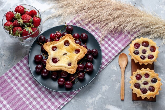 Vista superior de pequeños pasteles con frutas y crema junto con cerezas frescas fresas en mesa de luz pastel crema fruta té dulce