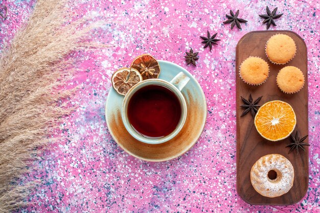 Vista superior de pequeños pasteles con rodaja de naranja y taza de té en superficie rosa claro