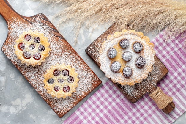 Vista superior pequeños pasteles sabrosos con frutas y azúcar en polvo en el escritorio blanco pastel galleta azúcar dulce