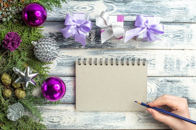 Vista superior pequeños regalos árbol de navidad juguetes ramas de abeto cuaderno lápiz en mano femenina sobre superficie de madera