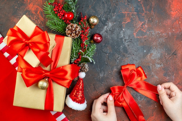 Vista superior pequeños regalos atados con cinta roja santa hat rama de árbol de navidad lazo rojo en mano femenina en mesa rojo oscuro
