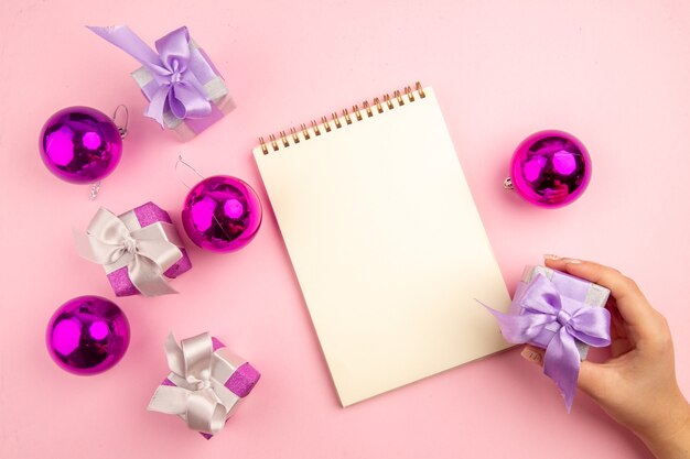 Vista superior de pequeños regalos con juguetes de árbol de Navidad y bloc de notas en la superficie rosa