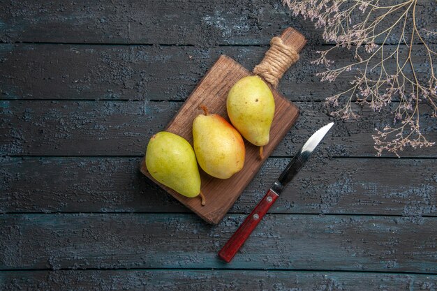 Vista superior de las peras y el cuchillo tres peras verdes, amarillas y rojas en el tablero de la cocina en el centro de la mesa oscura junto al cuchillo y las ramas de los árboles