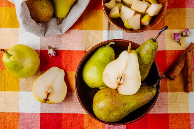 Vista superior de peras maduras frescas en un tazón de madera y mitades de pera con palitos de canela sobre fondo de cuadros