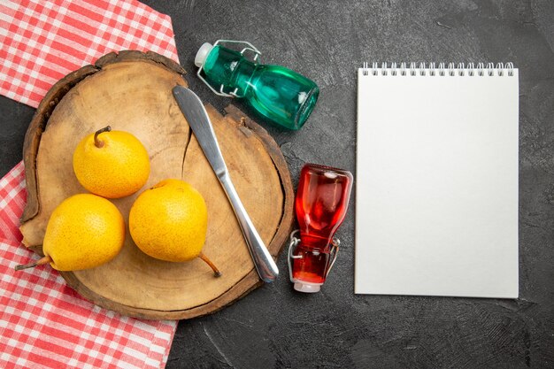 Vista superior de las peras en el tablero las apetitosas peras y un cuchillo en el tablero de la cocina sobre el mantel junto al cuaderno blanco y las botellas