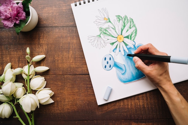 Vista superior de una persona dibujando un jarrón de flores sobre lienzo con un marcador negro en la mesa de madera