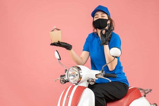 Vista superior de la persona de entrega feliz con máscara médica y guantes sentado en scooter entregando pedidos sobre fondo de melocotón pastel