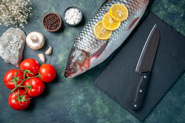 Vista superior de pescado crudo fresco con rodajas de limón y tomates sobre fondo azul oscuro