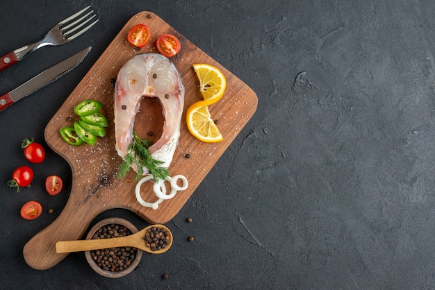 Vista superior de pescado crudo y verduras frescas picadas, rodajas de limón, especias en una tabla de madera, cubiertos en el lado derecho sobre una superficie angustiada negra