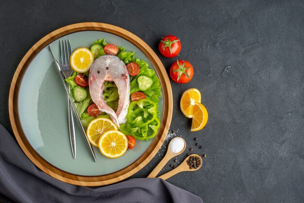 Vista superior de pescado crudo y verduras frescas, rodajas de limón y cubiertos en una placa gris, especias, toalla de color oscuro en el lado izquierdo sobre una superficie negra