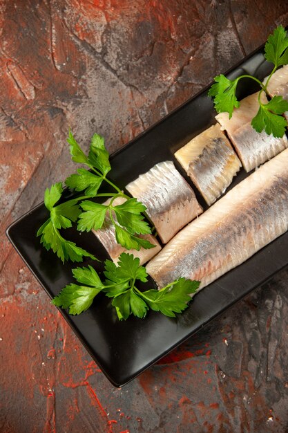 Vista superior de pescado fresco en rodajas con verduras y trozos de limón dentro de una sartén negra sobre el fondo oscuro