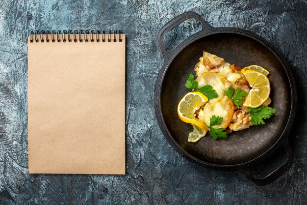 Vista superior de pescado frito en sartén con cuaderno de limón y perejil sobre fondo gris