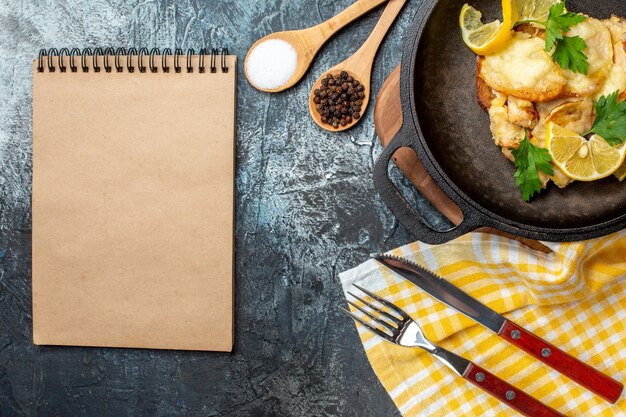 Vista superior de pescado frito en una sartén con especias de limón y perejil en un tazón y cucharas de madera, tenedor y cuchillo Bloc de notas sobre fondo gris