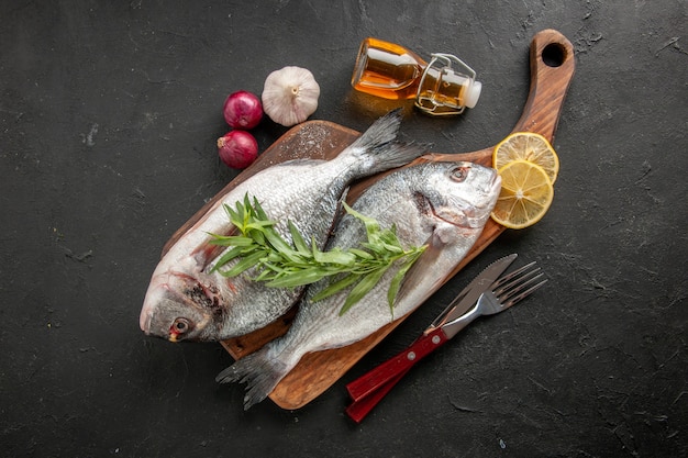 Vista superior de pescado de mar crudo en una tabla de cortar, tenedor y cuchillo, botella de aceite, cebollas gralic en negro