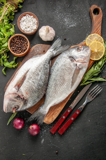 Vista superior de pescado de mar crudo en una tabla de cortar, tenedor y cuchillo, cebollas, sal marina y pimienta negra en tazones pequeños, menta sobre negro