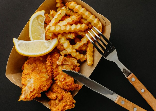 Vista superior de pescado y patatas fritas con cubertería y rodajas de limón