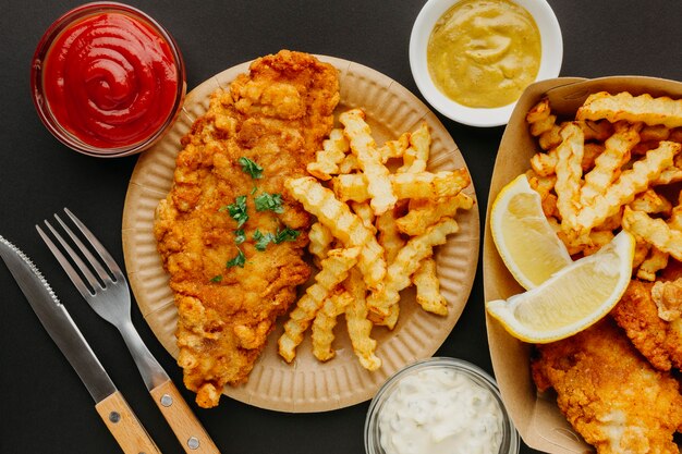 Vista superior de pescado y patatas fritas con cubiertos y selección de salsas