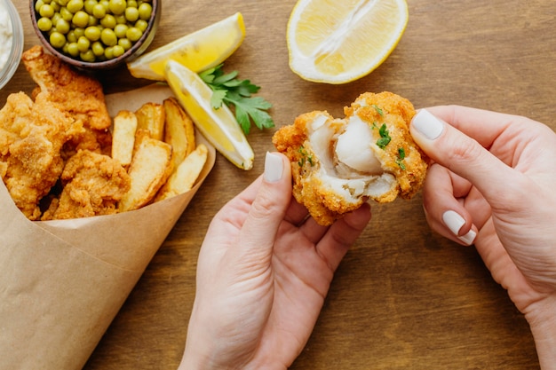 Vista superior de pescado y patatas fritas en papel con mujer rompiendo un trozo