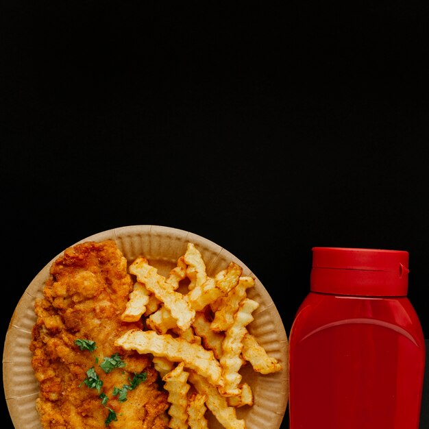 Vista superior de pescado y patatas fritas en un plato con botella de salsa de tomate y espacio de copia