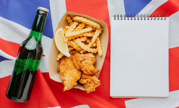 Vista superior de pescado y patatas fritas en un plato con un cuaderno y una botella de cerveza