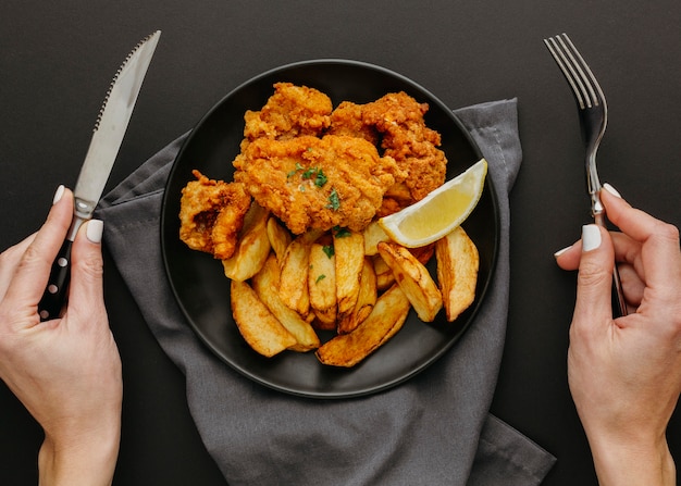 Vista superior de pescado y patatas fritas en un plato con mujer sosteniendo cubiertos