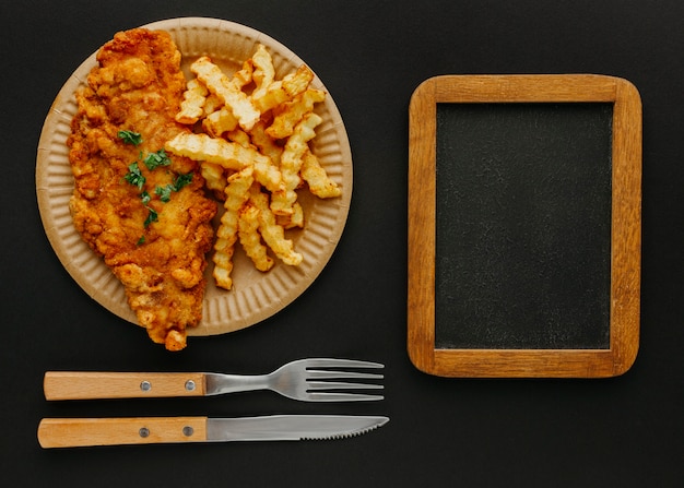 Vista superior de pescado y patatas fritas en un plato con pizarra y cubiertos