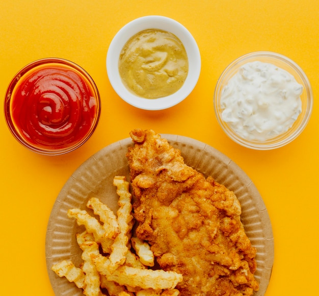 Vista superior de pescado y patatas fritas en un plato con salsas