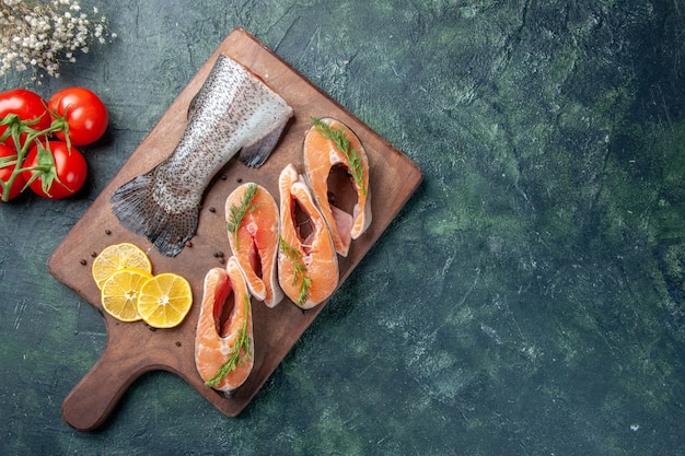 Vista superior de los pescados crudos, rodajas de limón, pimiento verde en tomates de tabla de cortar de madera en la mesa oscura