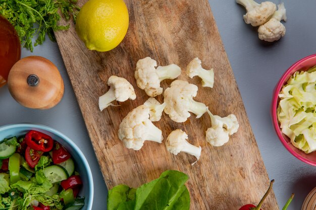 Vista superior de piezas de coliflor con espinacas y limón en la tabla de cortar con rodajas de col ensalada de sal y verduras de cilantro sobre fondo azul