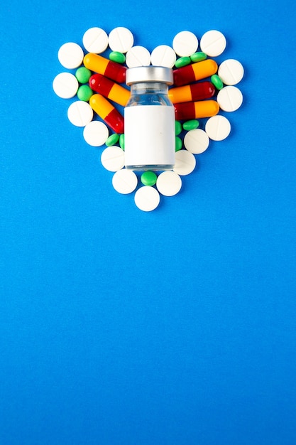 Vista superior de píldoras en forma de corazón de diferentes colores con vacuna sobre fondo azul.