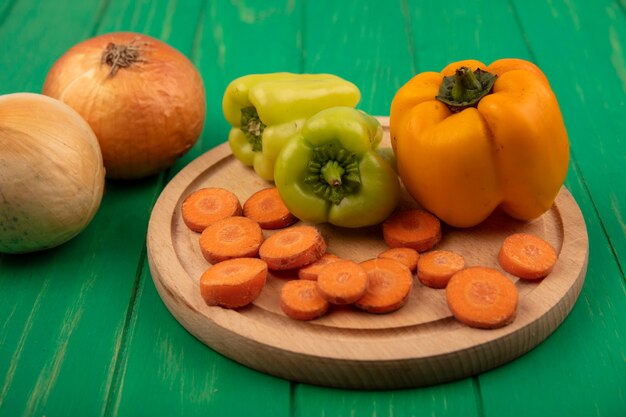 Vista superior de pimientos amarillos y verdes en una tabla de cocina de madera con zanahorias picadas con cebolla aislado en una pared de madera verde