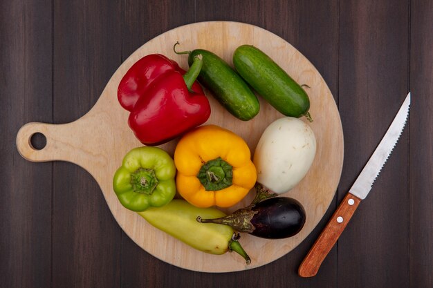 Vista superior de pimientos de colores con bklazana y pepino en una tabla de cortar con un cuchillo sobre un fondo de madera