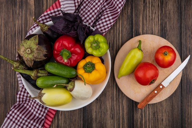 Vista superior de pimientos de colores con pepinos berenjena y baslik en un plato con tomates y un cuchillo sobre una tabla de cortar sobre un fondo de madera